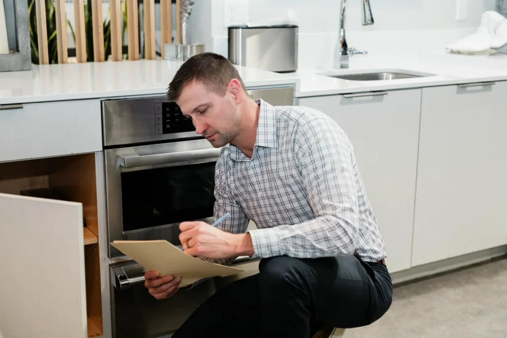 A man in business attire kneels in front of an open built-in oven, looking at a document in a modern kitchen.