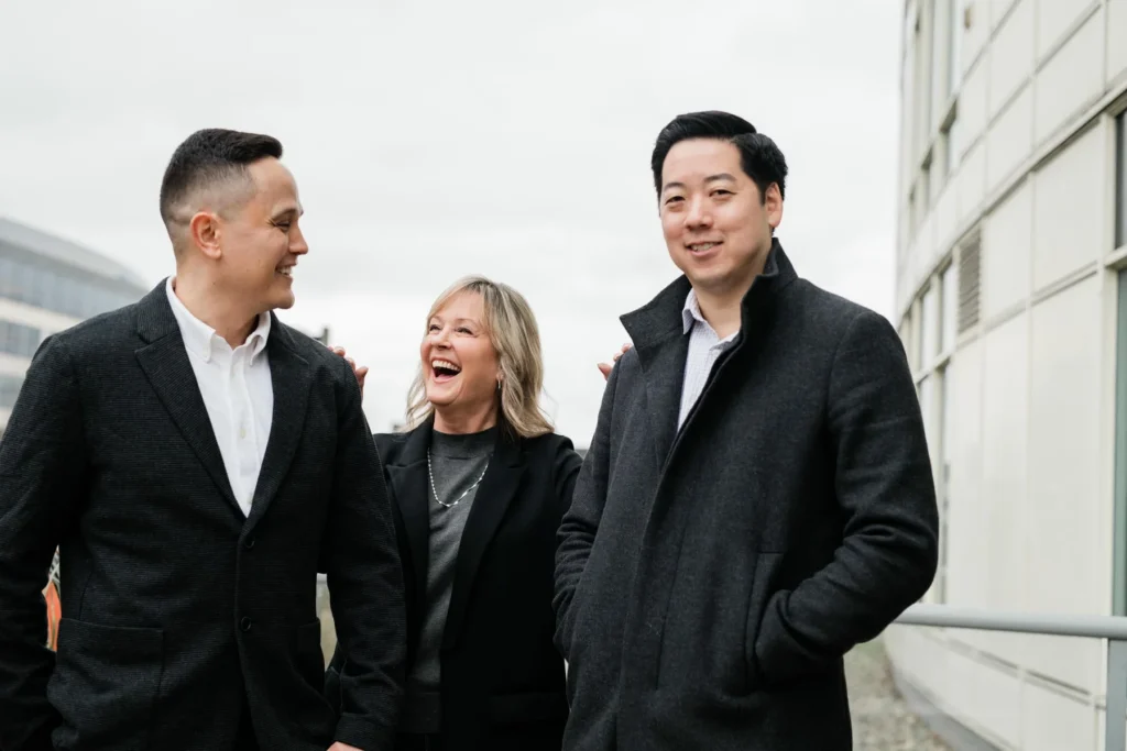 Three people in business attire stand outside a modern building. The woman in the middle is laughing, while the two men on either side look toward her and the camera.