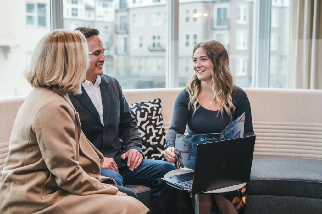 Three people sit on a couch in an office, talking and smiling, with a laptop and magazine open in front of them. Large windows show apartment buildings outside.