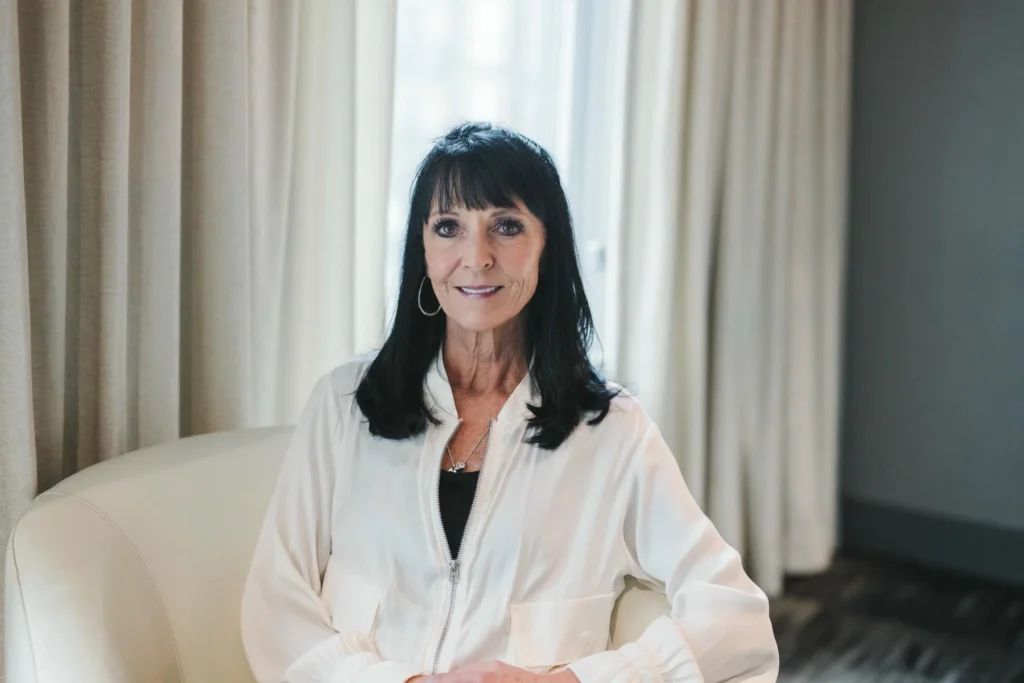A woman with straight dark hair and bangs sits on a light-colored chair, wearing a white zip-up top, in a room with light curtains and a window in the background.