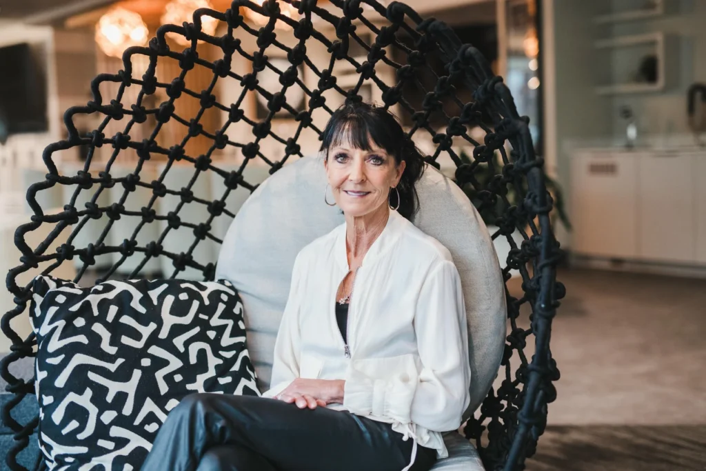 A woman with dark hair sits on a round woven chair, wearing a white blouse and black pants, with a patterned pillow beside her in a modern indoor setting.