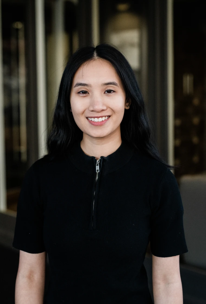 A woman with long black hair wearing a black zippered top stands indoors, smiling at the camera.
