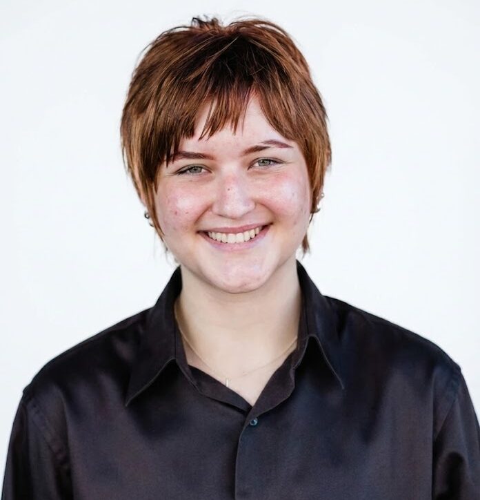 A person with short, light brown hair and a black collared shirt smiles at the camera against a plain white background.