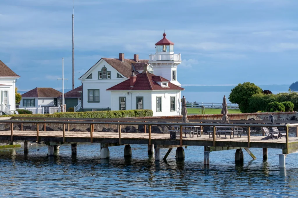 A white lighthouse with an attached house stands near the edge of a wooden pier, overlooking calm water under a partly cloudy sky.