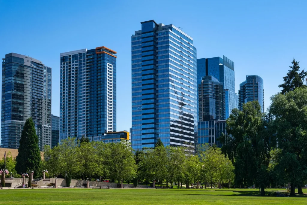 Modern high-rise office and residential buildings seen behind a park with green grass and trees under a clear blue sky.