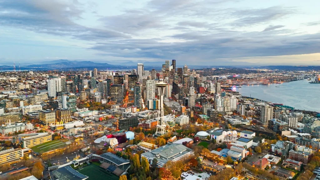 Aerial view of a modern city skyline with tall buildings, the Space Needle, green spaces, and water under a cloudy sky at sunset.