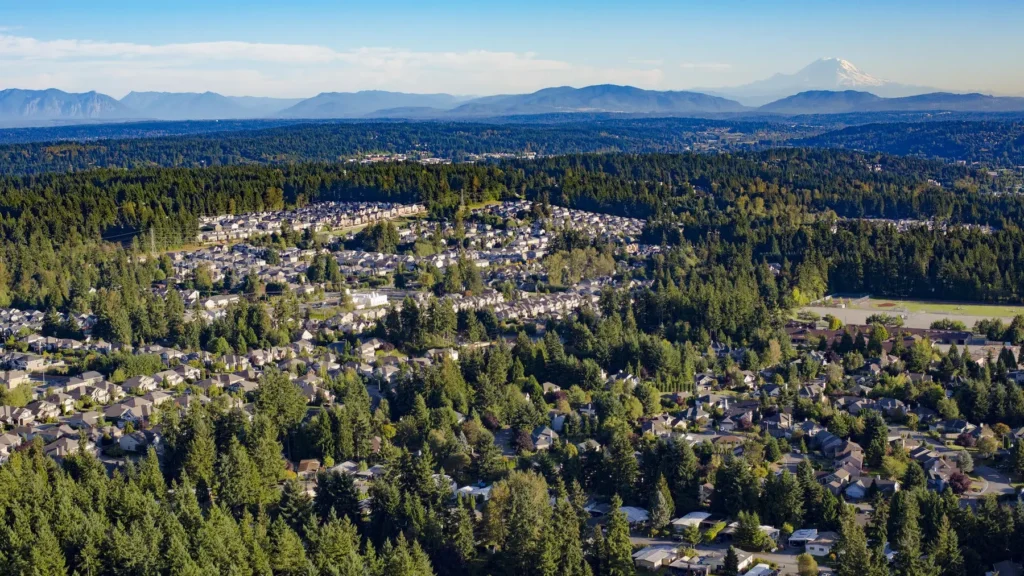 Aerial view of a suburban neighborhood surrounded by dense forest, with mountains visible in the background under a clear blue sky.