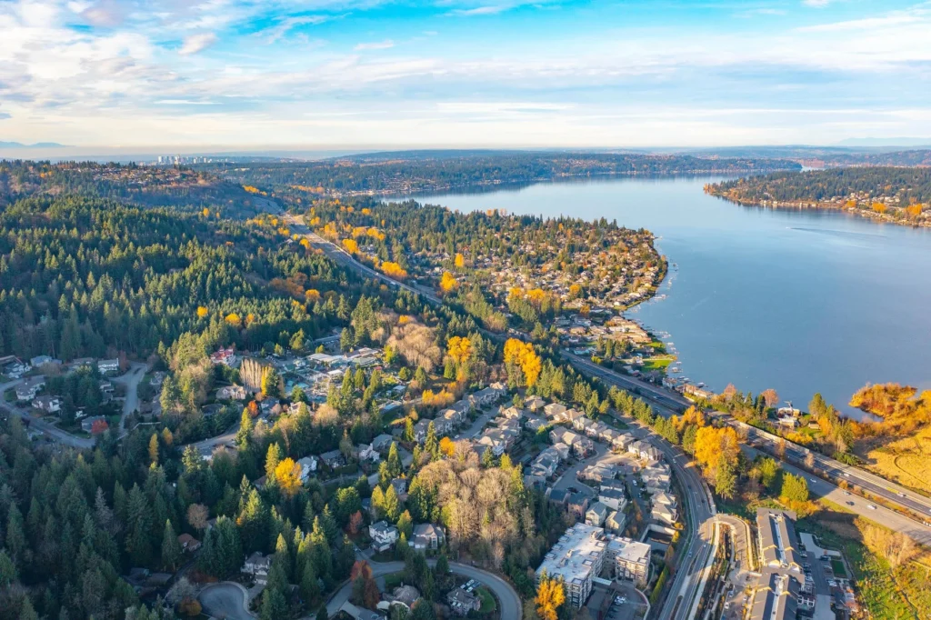 Aerial view of a suburban neighborhood with houses, dense trees, and a large lake, under a mostly clear sky. Roads and autumn-colored foliage are visible throughout the landscape.
