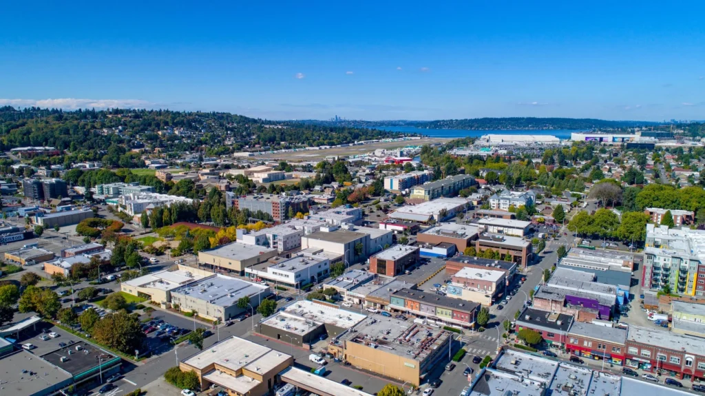 Aerial view of an urban neighborhood with commercial and residential buildings, tree-lined streets, and a hill and body of water in the background under a clear blue sky.