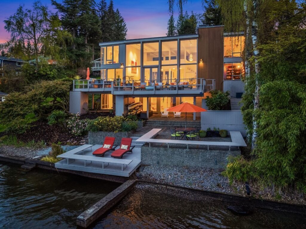 Modern lakeside house with large glass windows, outdoor seating areas, and a dock, surrounded by trees at sunset.