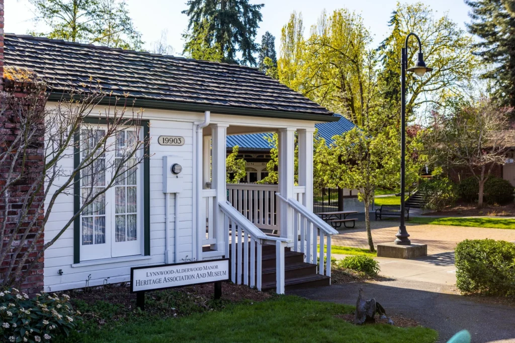 A white wooden building with a covered porch displays a sign reading “Lynnwood-Alderwood Manor Heritage Association & Museum” beside a walkway and trees.