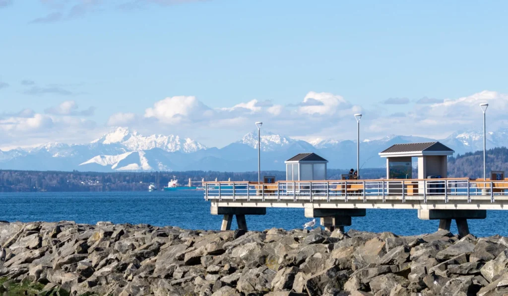 A pier with two small shelters extends over the water with snow-capped mountains and a partly cloudy sky in the background. Large rocks line the shore in the foreground.