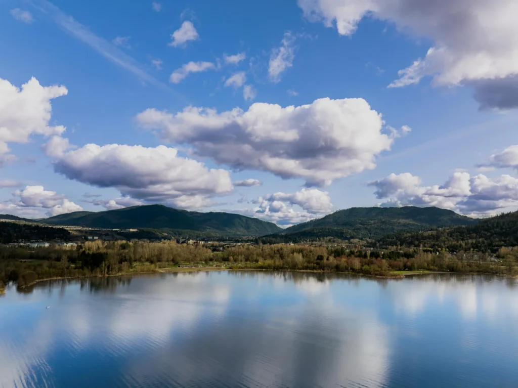 A calm lake reflects clouds and blue sky, with forested hills and scattered trees in the background under partly cloudy weather.