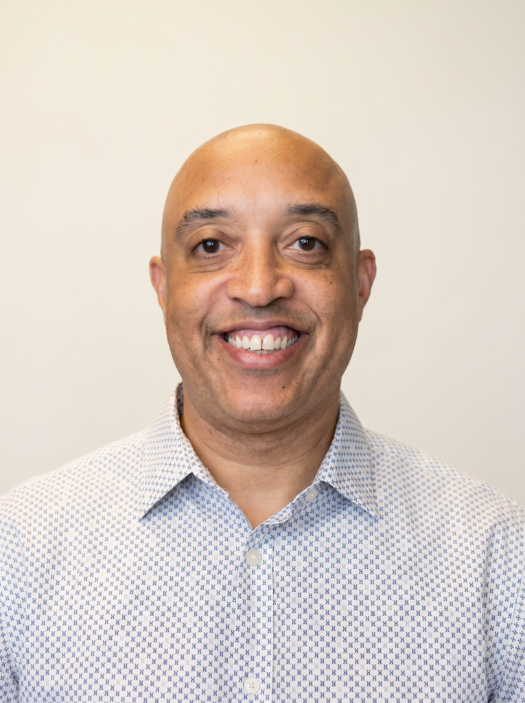A man with a shaved head smiles at the camera, wearing a patterned button-down shirt against a plain light background.