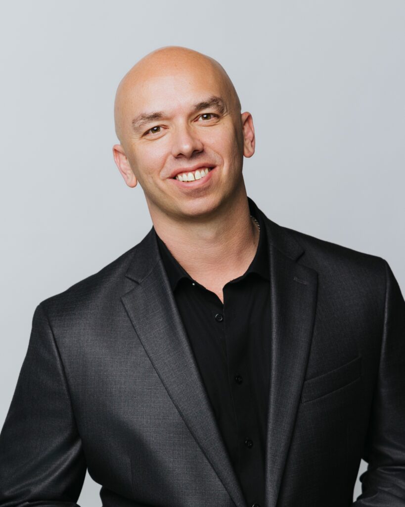 A smiling bald man wearing a black suit jacket and black shirt poses against a plain light gray background.