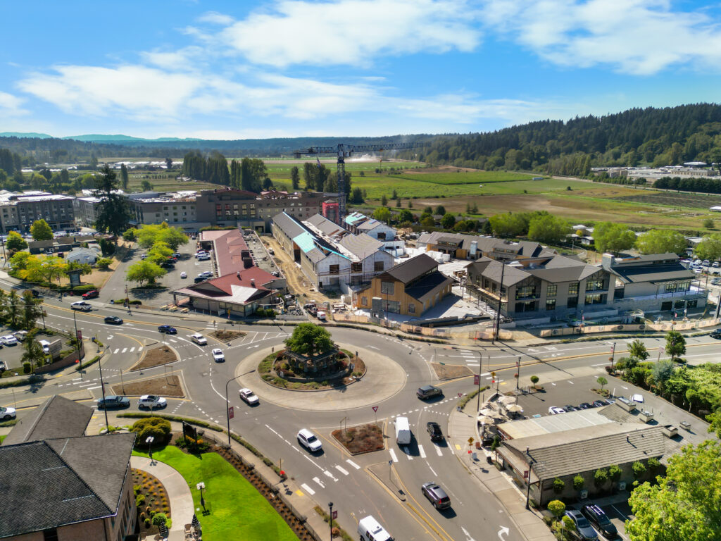 Aerial view of a roundabout surrounded by buildings, cars, and green fields under a partly cloudy sky.