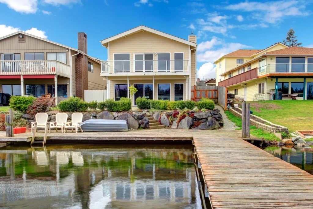 Waterfront house with large windows and a balcony, facing a wooden dock with outdoor chairs and a small hot tub by the water under a partly cloudy sky.