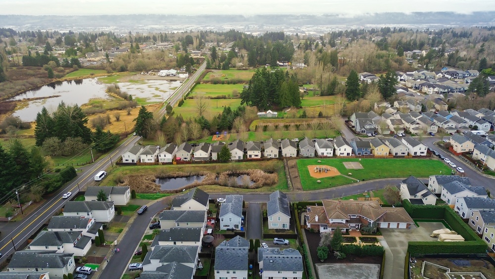 Aerial view of a suburban neighborhood with houses, a playground, small ponds, a road, trees, and open green spaces on a cloudy day.