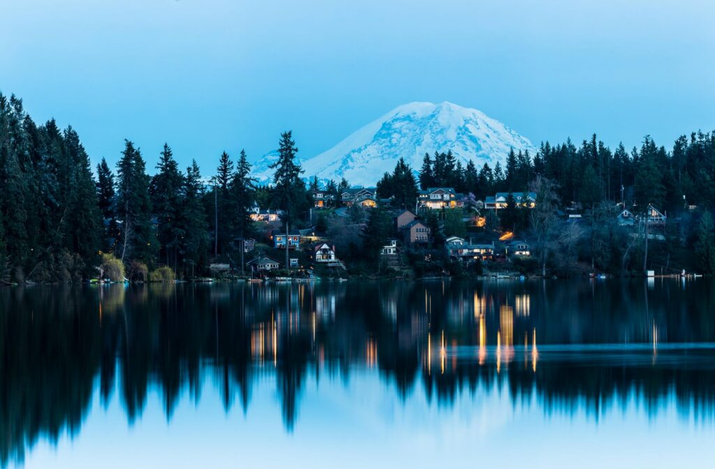 Snow-covered mountain rises behind a forested lakeshore with houses; evening lights reflect on the calm water.